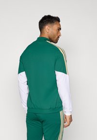 Zelená a biela športová súprava s bundou, so zlatými pruhmi na pleciach, rebrovanými manžetami a vysokým golierom. Hladká textura a priliehavý dizajn.