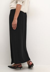 Jupe maxi noire fluide en tissu lisse, avec une coupe droite et de légentes fentes latérales. Associée à des escarpins à petits talons noirs.