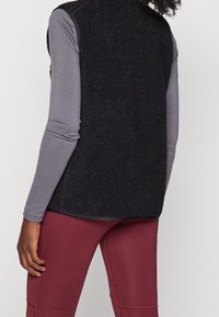 Black fleece vest with a textured surface, sleeveless design, and contrasting gray long-sleeve top beneath. Worn with maroon leggings.