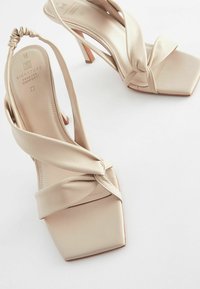 Beige High Heels Sandalen mit einem geknoteten Lederdesign, quadratische Zehen und einem schmalen Absatz. Glatte Textur und minimalistische Akzente.