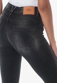 Los vaqueros de mezclilla negra tienen una cintura alta, costuras detalladas y un parche de cuero en la parte posterior. Se puede ver el diseño del bolsillo trasero.