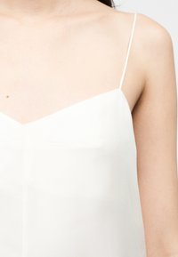 Γυναίκα με λευκό αμάνικο τοπ με λεπτούς ιμάντες, που δείχνει το πάνω μέρος του στήθους και τον δεξί ώμο σε ανοιχτό φωτισμό.