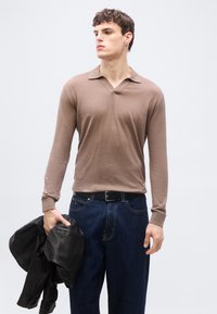 Jeune homme portant un polo à manches longues marron et un jean foncé, tenant une veste en cuir noire dans sa main gauche.