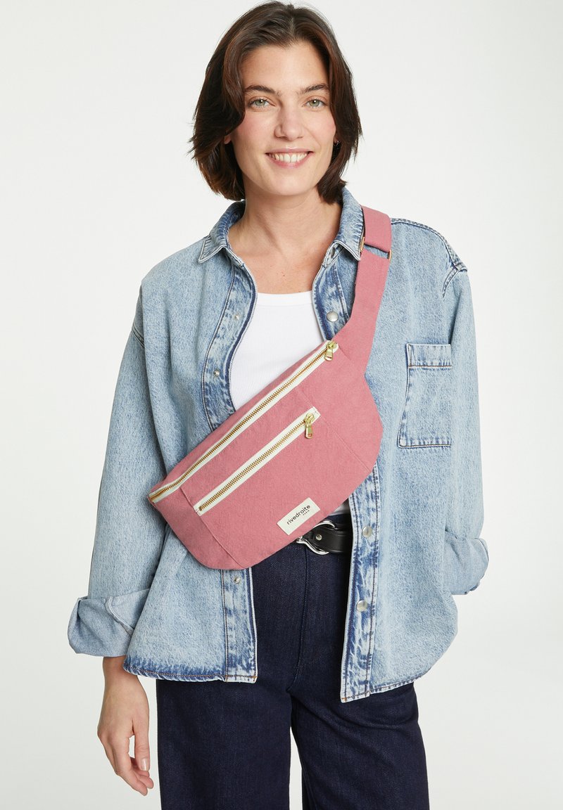 Pochette banane en tissu rose avec deux zips dorés, sangle attachée, et une étiquette. Le modèle porte une veste en jean sur un haut blanc.