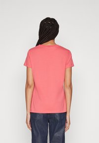 Kvinna med långt flätat hår, iklädd en enkel korallfärgad t-shirt med kort ärm och mörkblå jeans, står mot en vit bakgrund, sedd bakifrån.