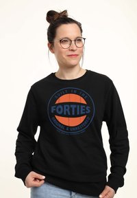 Mujer con gafas que lleva una sudadera negra con un gráfico naranja y azul que dice "Construido en los años cuarenta, original y sin restaurar."