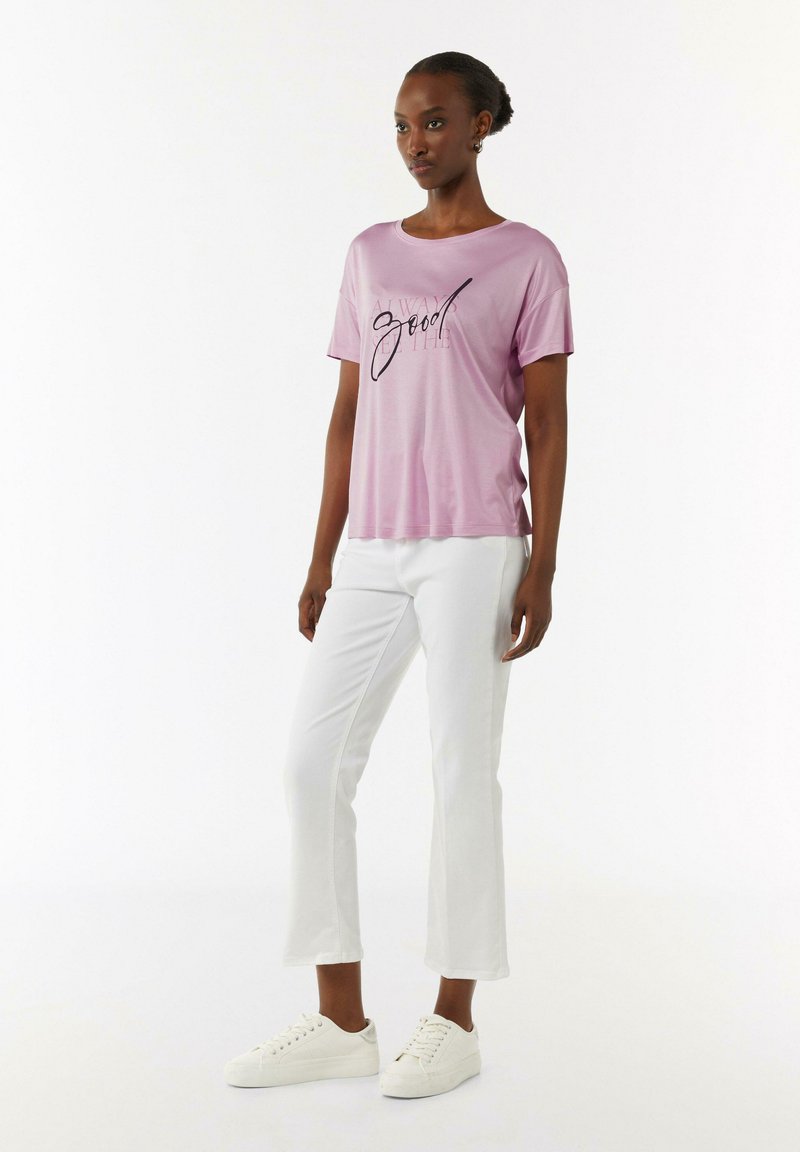Pink cotton t-shirt with short sleeves and a round neckline, featuring a black graphic text print, paired with white cropped pants and white sneakers.