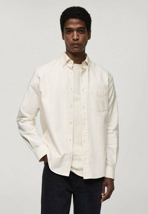 Light cream button-up shirt with a chest pocket, worn over a matching short-sleeve top. Features a classic collar and long sleeves.