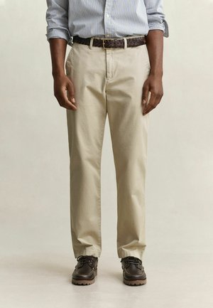 Homme portant un pantalon beige, une chemise à rayures boutonnée avec les manches retroussées, une ceinture tressée marron et des chaussures en cuir brun foncé.