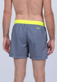 Maillots de bain en bleu marine avec un motif géométrique, dotés d'une ceinture élastique jaune vif et de poches latérales. Design arrivant au milieu de la cuisse.