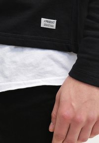 Schwarzes Langarmshirt, das über einem weißen Shirt getragen wird. Hat ein Stoffetikett mit "#PRODUKT INDUSTRIELL HERRGESTELLT" nahe dem Saum. Glatte Textur.