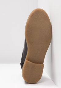 Semelle de chaussure avec une semelle en caoutchouc lisse marron clair, partie supérieure noire partiellement visible, positionnée à la verticale contre une surface blanche en angle.