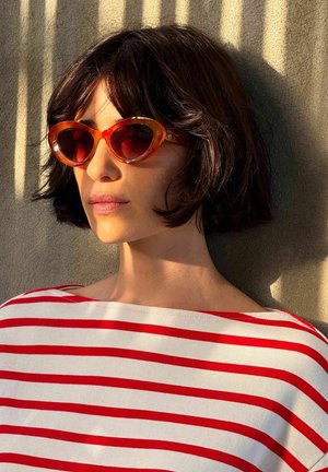 Vrouw met kort donker haar, draagster van oranje hartvormige zonnebril en een rood-wit gestreept shirt, verlicht door zonlicht en schaduwen.