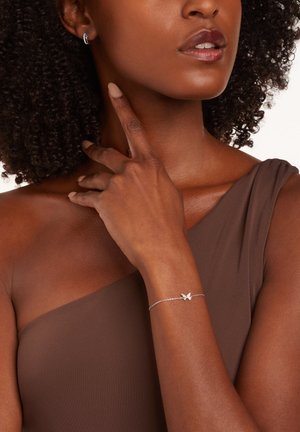 Bracelet en argent en forme de papillon avec une fine chaîne ; porté au poignet. Le teint de la peau contraste avec le top marron à une épaule et le fond texturé.