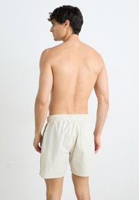 Shorts de bain couleur crème avec une taille élastique et une poche à l'arrière, fabriqués à partir d'un matériau léger, présentant un design simple et minimal.