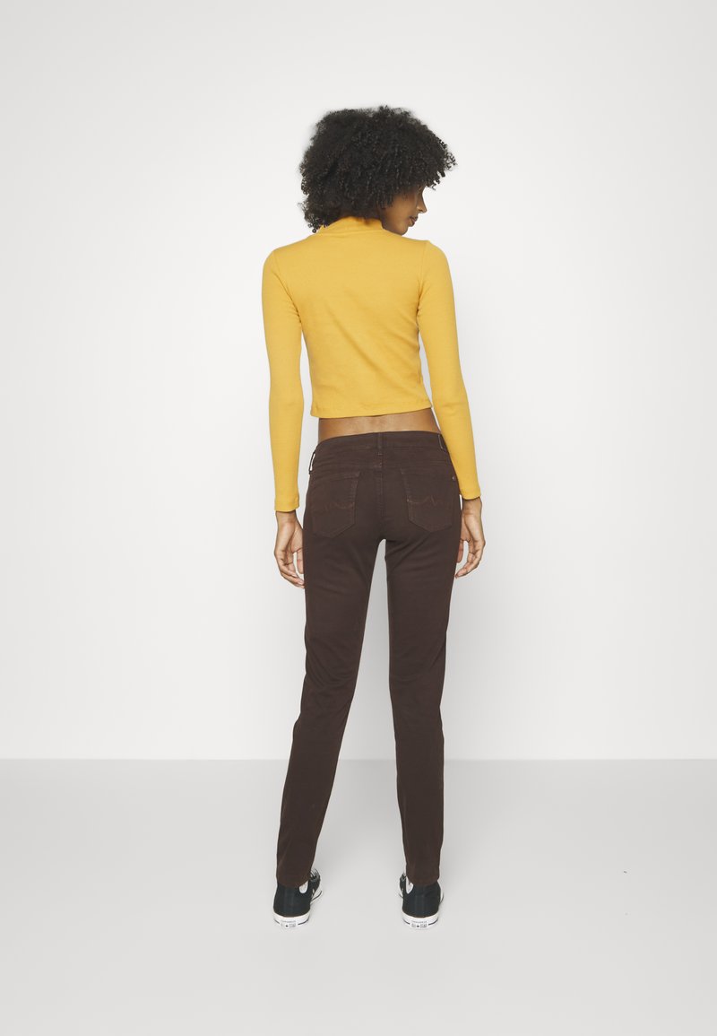 Braune, taillierte Hose mit hoher Taille und aufgesetzten Taschen, kombiniert mit einem kurzgeschnittenen, senfgelben Langarmoberteil. Schwarze Turnschuhe runden das Outfit ab.