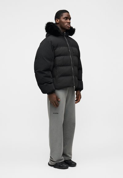 Casaco acolchoado preto com capuz forrado a pelo, com fecho de correr e punhos elásticos. Usado com calças de fato de treino cinzentas e calçado preto.