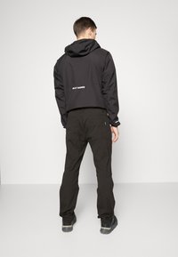 Crna vodootporna jakna s kapuljačom, koja ima skraćeni dizajn i vidljiv logotip na leđima. Uparena s labavim crnim hlačama.