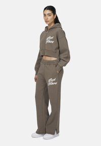Hoodie marrone cropped con testo "Good for Nothing", abbinato a pantaloni marroni coordinati con spacchi laterali, realizzati in tessuto morbido e testurizzato.