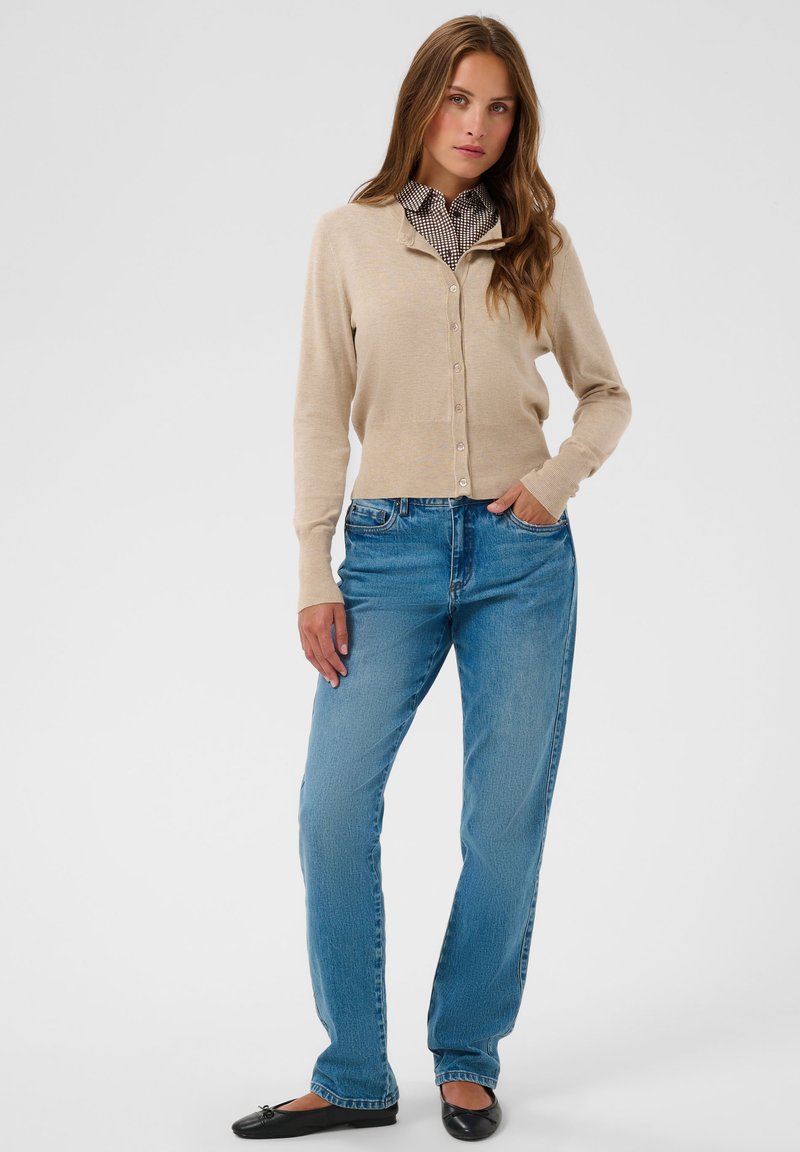 Femme portant un cardigan beige boutonné sur une chemise à carreaux, un jean bleu et des ballerines noires, debout avec une main dans la poche devant un fond blanc.