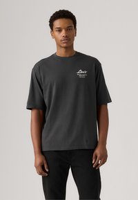 Camiseta gris de manga corta hecha de algodón con un cuello redondo. Presenta un logotipo impreso en blanco en la zona del pecho izquierdo. Textura suave.