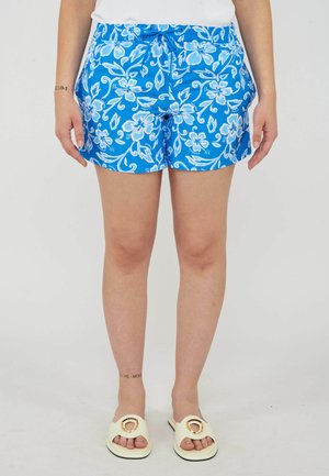 Persona che indossa pantaloncini blu a fantasia floreale, top bianco e sandali crema, in piedi davanti a uno sfondo semplice.