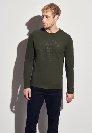 Camicia verde oliva a maniche lunghe con dettaglio stampato texturizzato sul petto, abbinata a jeans scuri, presentata su uno sfondo neutro.