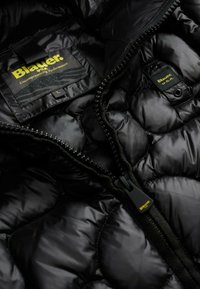 Interno di giacca a vento nera con una texture lucida, zip visibili e etichette "Blauer" in giallo e verde. Presenta un design trapuntato.