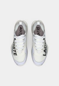 Λευκά αθλητικά παπούτσια με υφασμάτινο πάνω μέρος με υφή, έντονη μαύρη σήμανση "LACOSTE" στην πλευρά και μαξιλαρωτή σόλα.