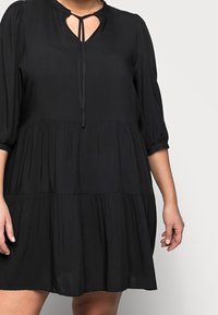 Vestido negro hecho de tejido ligero, con escote en V con lazo, mangas 3/4 y un diseño de volantes y fluido. La textura es suave.