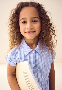 Mėlynos ir baltos spalvos languota trumpomis rankovėmis suknelė su apykakle; laiko kremiškai baltos spalvos aplanką su subtiliomis linijomis. Minkšti, garbanoti plaukai rėminantys veidą.