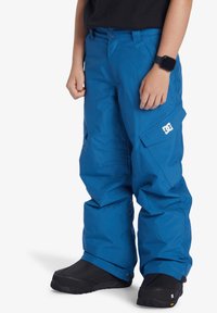 Blå snöbyxor tillverkade av vattentåligt material, med en logga, sidofickor och en lös passform. Ett par svarta vinterstövlar syns nedanför.