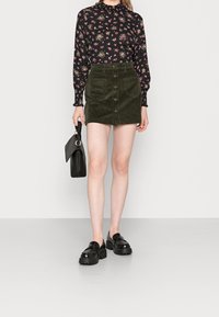 Blusa nera a fiori con maniche lunghe, gonna in velluto a coste verde con bottoni frontali, borsa nera con manico intrecciato e scarpe nere chunky.
