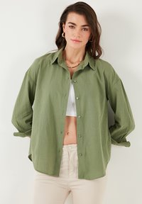 Olive green, button-up shirt with a collar, long sleeves, and a relaxed fit. Lightweight fabric with a smooth texture, worn over a white tank.