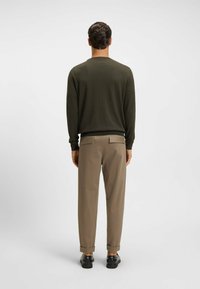 Maglione verde oliva con collo e polsini a coste, abbinato a pantaloni marrone chiaro leggermente arrotolati all'orlo, indossato con scarpe nere.
