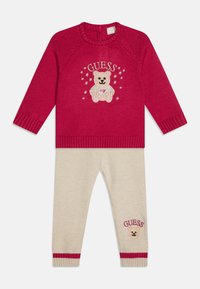 Maglione rosso lavorato a maglia con ricamo di orsetto e stelle, abbinato a pantaloni beige con orlo a righe rosse e un dettaglio di orsetto coordinato.