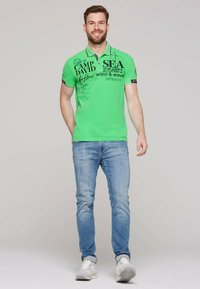 Zielona koszulka polo z czarnym graficznym napisem i kołnierzykiem, w połączeniu z jasnoniebieskimi dżinsami i białymi trampkami. Gładka tekstura i casualowy styl.