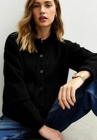 Zwarte wollen cardigan met ronde halslijn, knopen aan de voorkant en een gestructureerde afwerking. Gecombineerd met blauwe denim jeans en een gouden horloge.
