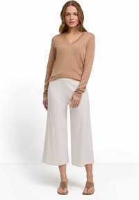 Beżowy sweter w serek zestawiony z białymi, prążkowanymi, szerokimi spodniami. Sweter ma długie rękawy; obuwie to jasnobrązowe klapki.