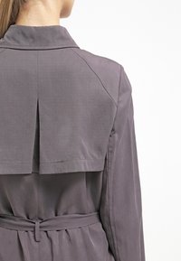 Szary płaszcz typu trench, z plisą na plecach, usztywnionym kołnierzem i paskiem w talii. Miła w dotyku struktura z gładkim, lekko błyszczącym wykończeniem.