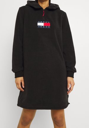 Osoba ubrana w czarną, długorękawową sukienkę z kapturem, zapinaną na przedni zamek błyskawiczny, z naszywką z logo Tommy Jeans na piersi.