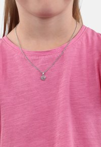 Silverhalsband med ett hjärtformat hänge som har flera rosa stenar, bärs över en ljusrosa, texturerad T-shirt.