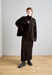 Černá džínová bunda se stříbrnými knoflíky, přiléhavý černý ribbed top s výřezem, dlouhá černá sukně a robustní černé kotníkové boty na dřevěné podlaze.