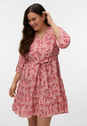 Ung kvinde smilende, iført en pink blomsterprintet V-hals kjole med 3/4 ærmer og et bundet bælte i taljen, der rører ved sit hår foran en ensfarvet baggrund.