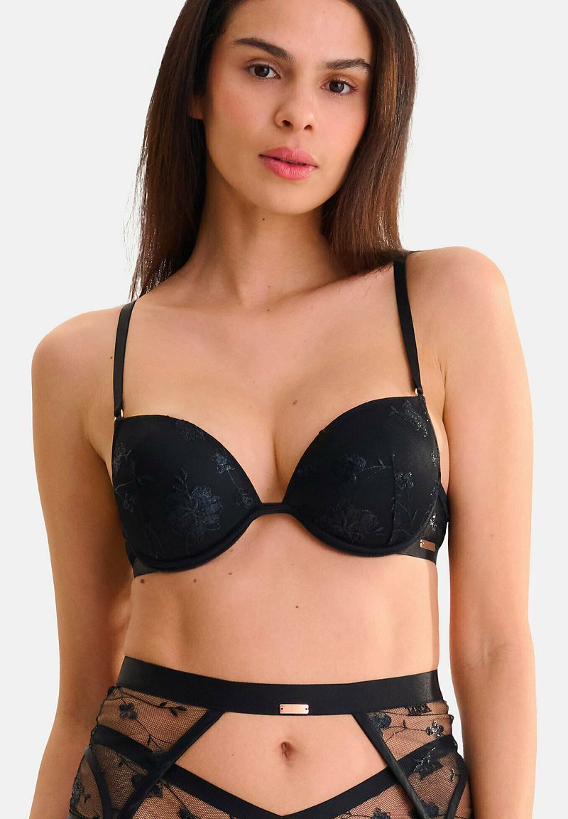 Soutien-gorge rembourré noir avec broderie florale, fines bretelles ajustables et texture lisse. Inclut un accent métal au centre de la bande sous la poitrine.