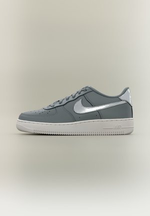 Baskets Nike Air Force 1 grises avec un swoosh argenté métallique et une languette au talon, semelle blanche, présentées sur fond neutre, vue de profil.