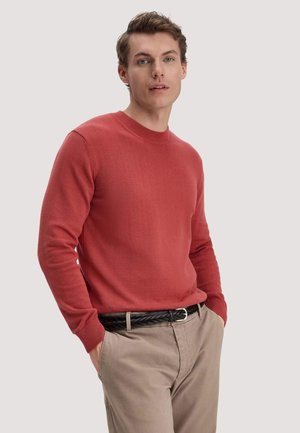 Junger Mann steht mit den Händen in den Taschen, trägt einen roten Pullover und beige Hose mit einem schwarzen geflochtenen Gürtel vor einem schlichten Hintergrund.