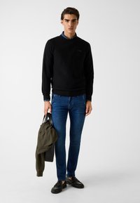 Schwarzer Pullover, blaue Hemd darunter, blaue Jeans, hält eine grüne Tasche. Die Schuhe sind schwarz mit Fransenverzierung. Einfaches Design und taillierte Form.