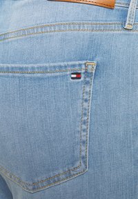 Jeans de mezclilla azul claro con un bolsillo trasero que incluye un pequeño logo en rojo, blanco y negro, y detalles de costuras amarillas en contraste.