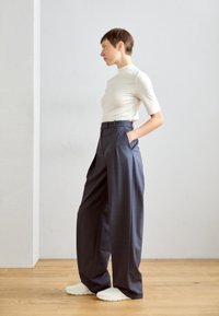 Ljust beige figurtopp, högmidjade mörkgrå vida byxor och vita slip-on skor, mot en ljusgrå vägg och trägolv.